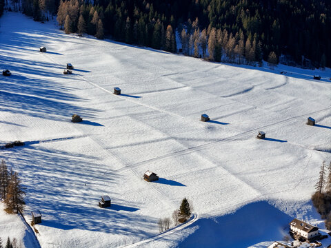 Aerial view of winter's embrace, where snow-covered fields meet the rustic charm of wooden barns, a serene landscape under a clear sky, Sexten, Trentino-South Tyrol, Italy.