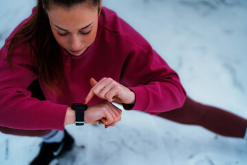Woman standing in snow and wearing activewear, checking her fitness smartwatch during a cold winter...