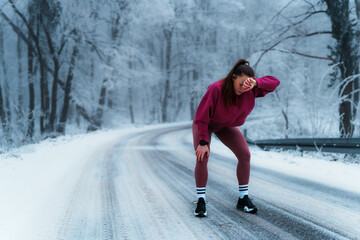 Young athletic woman wiping sweat and catching breath, taking a break from running on a snow covered road through a frozen forest park, showing determination and endurance in cold weather