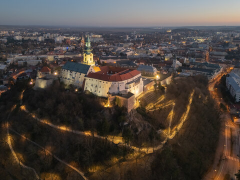 Aerial view of Nitra Castle perched atop a hill, illuminated pathways winding through the landscape in the twilight, Nitra, Nitriansky kraj, Slovakia.