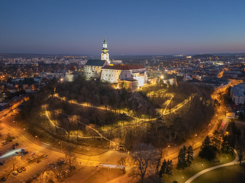 Aerial view of Nitra Castle perched atop a hill with winding paths illuminated against the twilight, Nitra, Nitriansky kraj, Slovakia.