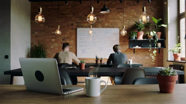 Steaming coffee mug and a laptop on a wooden desk with two male coworkers brainstorming in front of a whiteboard in the background of a modern loft style open space office or coworking area