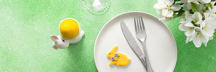 A beautifully arranged Easter table features a plate with a yellow bunny cookie, cutlery, and decorative elements like flowers and a small bunny figure, creating a festive atmosphere, banner