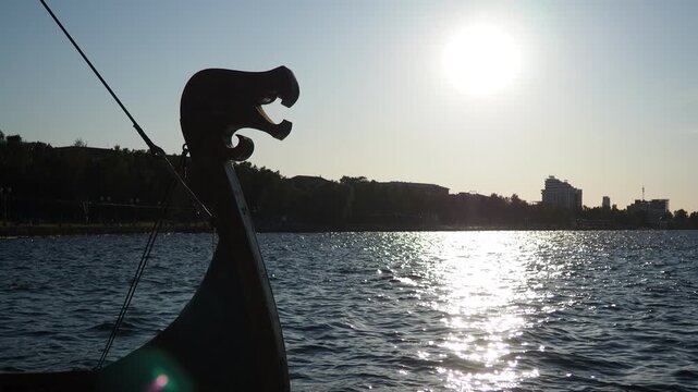 Petrozavodsk, Karelia, Russia. Wooden antique-style sailing boat sails in waters of Bay Lake Onega. Tourism in Summer sunny evening, blue waves. Carved wooden head of pagan totem, serpent or dragon