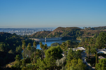 Obraz premium Los Angeles, USA - October 18, 2025 - Hollywood Reservoir dam and city skyline view with lush green trees and palm trees