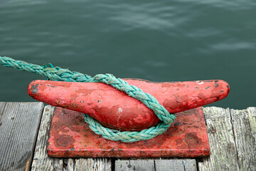Naklejka premium Close-up of a weathered red metal dock cleat wrapped with green rope on a wooden pier beside calm water. Nautical detail showing texture, wear, and marine elements, suitable for boating, maritime, har