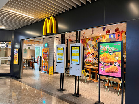 A McDonald's restaurant entrance showcasing a vibrant interior with colorful decorations and digital ordering kiosks. The iconic golden arches are prominently displayed.