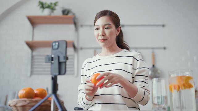 Young beautiful asian content creator recording a video or live streaming on her smartphone about healthy nutrition and infused water for longevity in a kitchen.