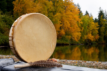 A close up image of a hand made leather meditation drum with a sacred feather and colorful autumn leaves reflecting off clear lake waters in the background. 
