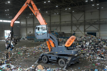 A large, specialized machine is actively sorting through heaps of household garbage in a waste management facility. The worker uses grapples to handle various materials, ensuring proper recycling