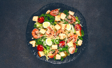 caesar salad with tiger prawns, shrimp salad, lettuce leaves, quail eggs, cherry tomatoes, parmesan cheese, on a black plate,