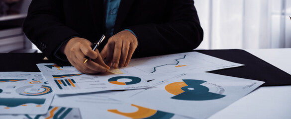 A focused business professional reviews financial documents and graphs, taking notes with a pen in a modern office setting, emphasizing strategic planning and analysis.Noogenesis