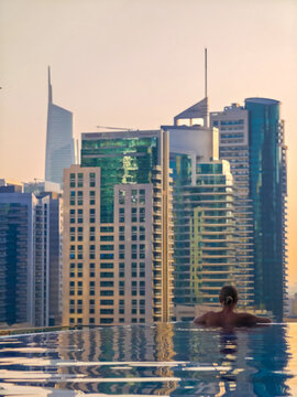 Woman relaxing in infinity pool at sunset, watching city skyscrapers, luxury lifestyle and urban skyline view. Dubai background