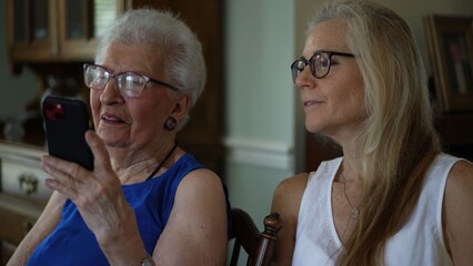 Senior woman and her daughter sit together looking at pictures and talking on a phone while sharing memories and moments