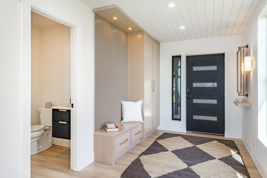 Entry foyer hallway area rug geometrical pattern hardwood flooring black front door large window blue sky cloak room coat hooks welcome bench arrival warm contemporary home interior designer decor
