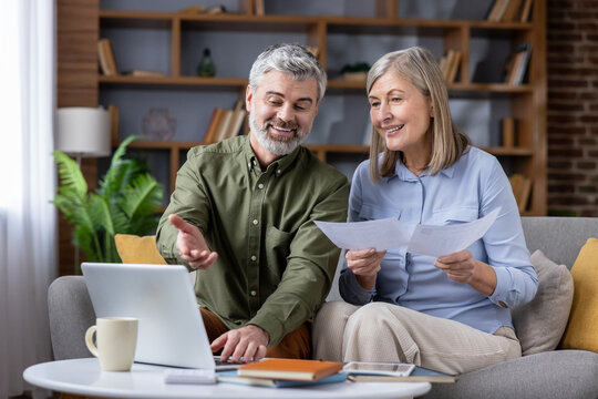 Senior couple sitting on a sofa discussing financial documents and managing household budget online, both smiling and looking at a laptop in their cozy living room at home - Powered by Adobe