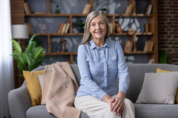 Senior woman with gray hair smiling confidently, sitting comfortably on a sofa in a modern cozy living room with a bookshelf in the background, representing aging gracefully and a happy lifestyle