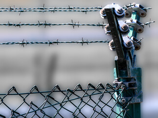 Close-up of a metal fence with barbed wire and green post, visible insulators and fixings, blurred background