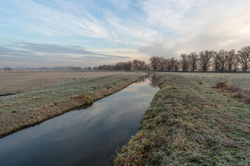 Frosty rural landscape with water canal at sunrise. Rural landscape with a narrow water canal running through frost-covered fields on a cold winter morning. 