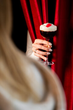 A person's hand holds a creamy, layered cocktail in a stemmed glass, garnished with a red heart and cherry, against dramatic red velvet curtains in a bar