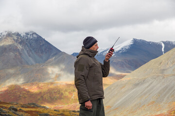 A European man talks on a radio against the backdrop of a mountain landscape.
