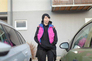Fototapeta premium Young person in bright vest standing between parked cars, carrying backpack and beanie, confident urban commuter ready for morning route, candid street style, cool color palette