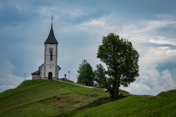 Iconic Church of Saints Primus and Felicianstanding on a scenic hilltop ridge under a dramatic cloudy sky with a gravel path  in Jamnik, Slovenia, 