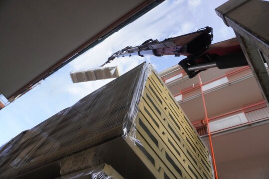 A construction crane lifts insulated roofing panels. A hydraulic truck crane precisely moves large, shrink-wrapped stacks of insulated sandwich panels between closely spaced residential buildings.
