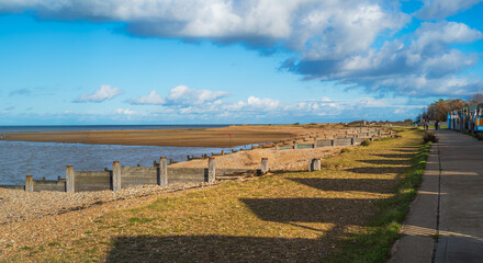 Whitstable beach panoramic scene. The beach is a pebble beach with wooden groynes. The day is bright in November with long shadows of the beach huts along the promenade.