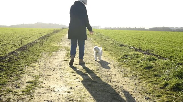 A woman in a black jacket walks a white Bichon Frise dog down a dirt path through a green field. They eventually turn around and run back toward the camera.