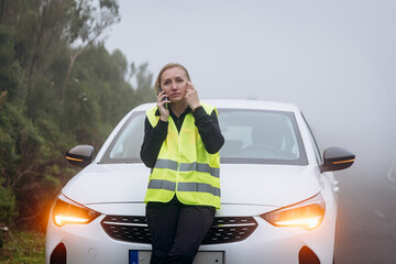 Woman in reflective vest is sitting on a car hood, talking on phone while experiencing vehicle trouble in foggy weather