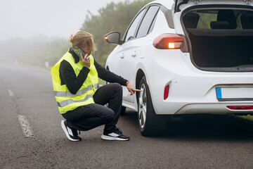 Woman in reflective vest checking car tire on roadside while talking on phone during foggy weather conditions