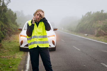 Woman in reflective vest experiencing car trouble on a foggy road while making a phone call for assistance