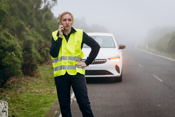 Woman in reflective vest standing by car on roadside, experiencing vehicle issues in foggy weather