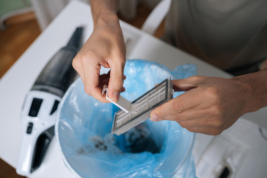 Person cleaning HEPA filter from dirty robot vacuum cleaner, carefully tapping out accumulated dust and debris into bin with blue plastic liner, performing essential device maintenance.