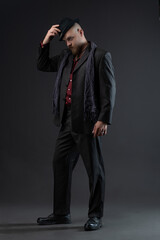 Studio portrait of charismatic male in noir suit accented with ethnic style details, complete with period hat, shot against neutral gray backdrop.