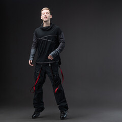 Young man in loose cyberpunk streetwear posing in studio on gray background.