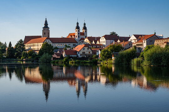 A wide panoramic view of the historic town of Telč with its colorful buildings and church towers reflected in the calm surface of a pond under a clear blue sky in the Czech Republic.