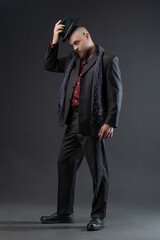 A man dressed in a noir style suit with elements of nonchalance and ethnic style, a charismatic man in a suit and hat in a photo studio on a gray background