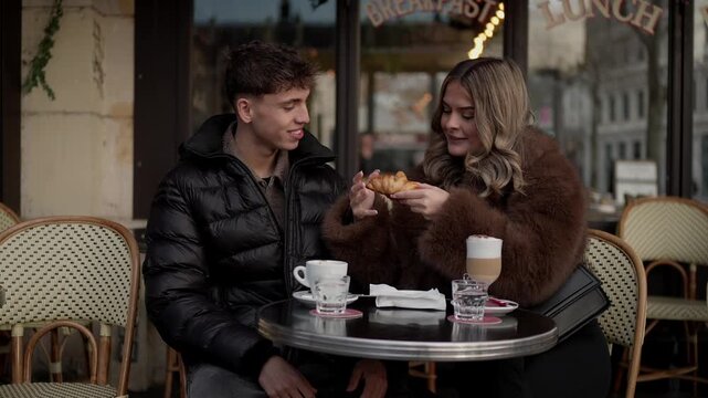 A young couple enjoys breakfast at an outdoor in Paris France The woman eats a croissant while the man looks on Coffee and water are on the table The scene captures a romantic moment in a European cit