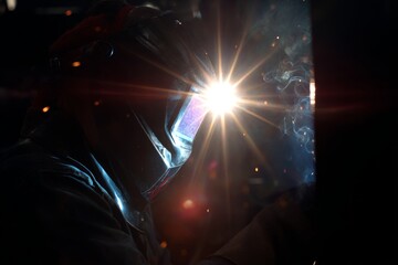 A welder wearing a protective helmet works with metal, producing a brilliant welding arc and flying sparks in a dark industrial setting.