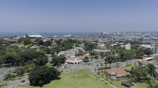 Drone lowers over Jameson Park with white stadium and CBD in the distance on sunny day in Morningside neighborhood in Durban, South Africa