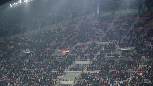 Crowd of sports fans supporters cheering during a football match in stadium. Excited people standing with their arms raised, clapping and yelling to encourage their team, slow motion