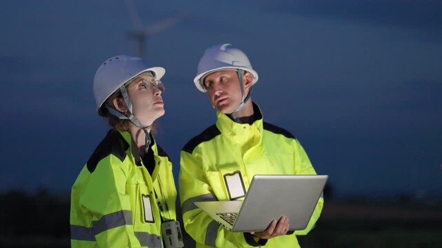 Two professional engineers in safety uniforms and hard hats working together with a laptop to monitor wind turbine performance at a renewable energy site during twilight.