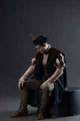 Fototapeta premium Cinematic romantic hero from medieval setting wearing rich brocade garments with flowing cape and period beret, dramatic posing on neutral gray backdrop