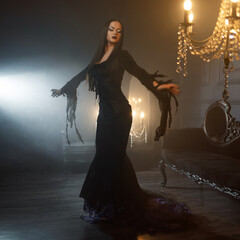 A gothic femme fatale with long straight hair and dark makeup wears a black godet skirt dress with corset, posed in a luxurious dimly lit gothic interior. © Ulia Koltyrina