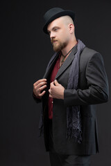 Fashionable man wearing a noir-inspired suit with ethnic decorative elements and matching hat, posing confidently in studio against simple gray background