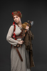 Nordic warrior woman in natural dress holding axe, fur cape on gray background.