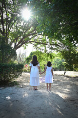 mother and daughter holding hands in sunny park, rear view of woman and girl in white dresses walking outdoors, family love and togetherness concept in nature, bright summer morning lifestyle