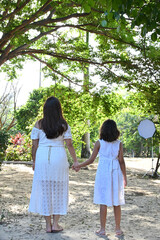 mother and daughter holding hands in sunny park, rear view of woman and girl in white dresses walking outdoors, family love and togetherness concept in nature, bright summer morning lifestyle